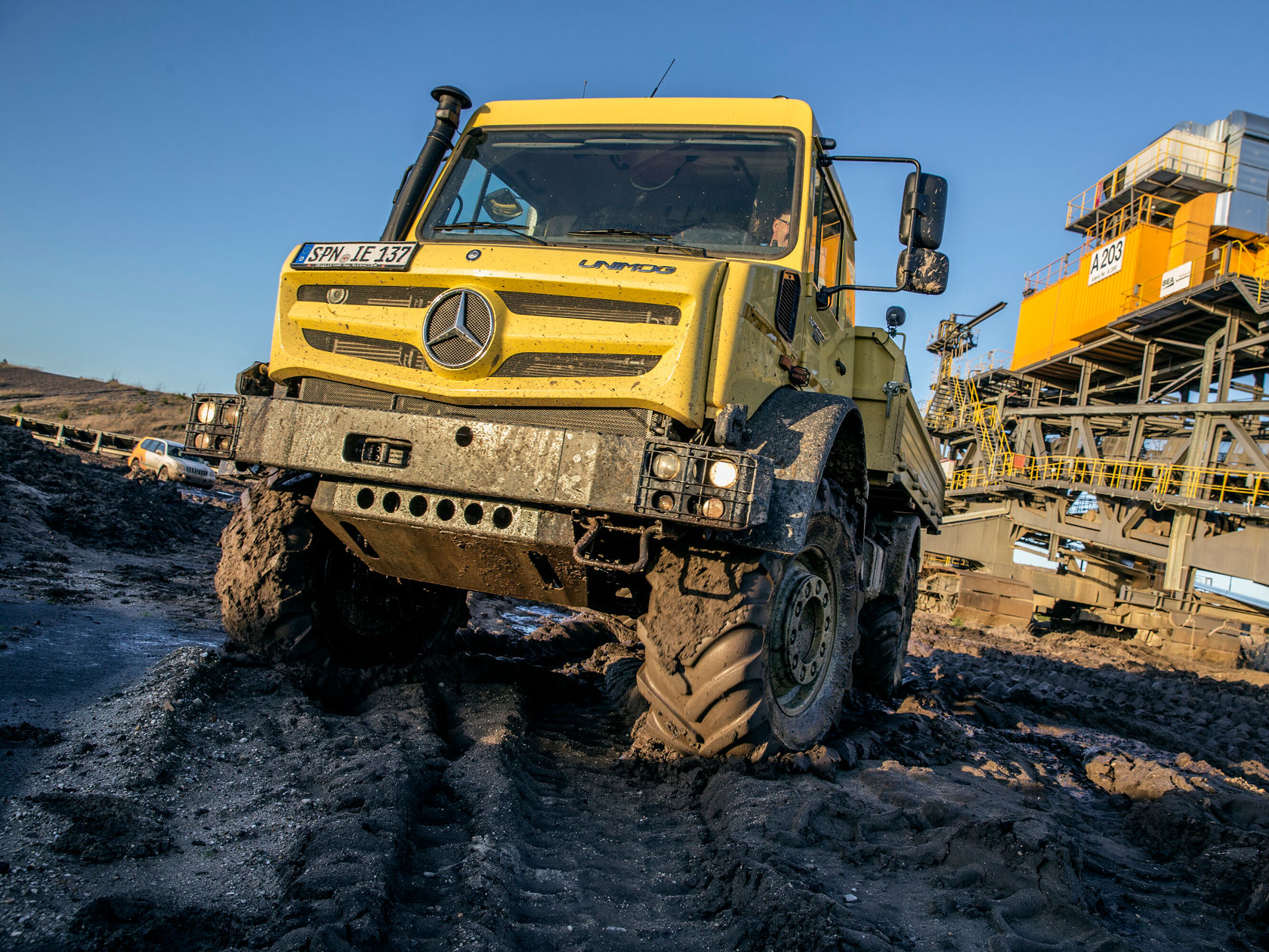 Un Unimog Mercedes-Benz jaune traverse un terrain boueux, tandis qu'un chantier est visible en arrière-plan. Le camion est conçu pour des missions exigeantes.