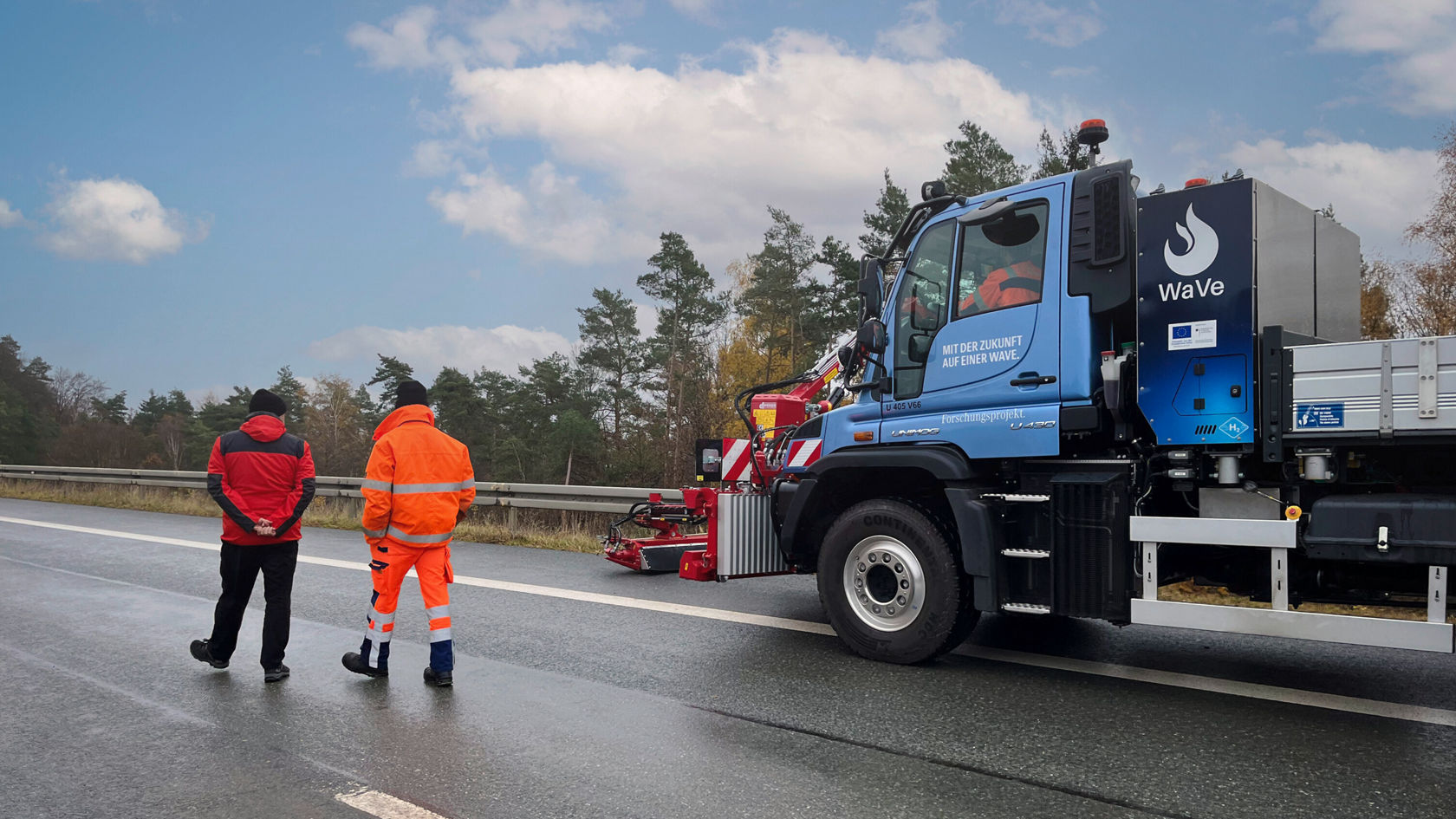 Deux personnes se tiennent au bord d'une autoroute à côté d'un Unimog Mercedes‑Benz équipé d'un réservoir d'eau, tandis qu'elles regardent la route.