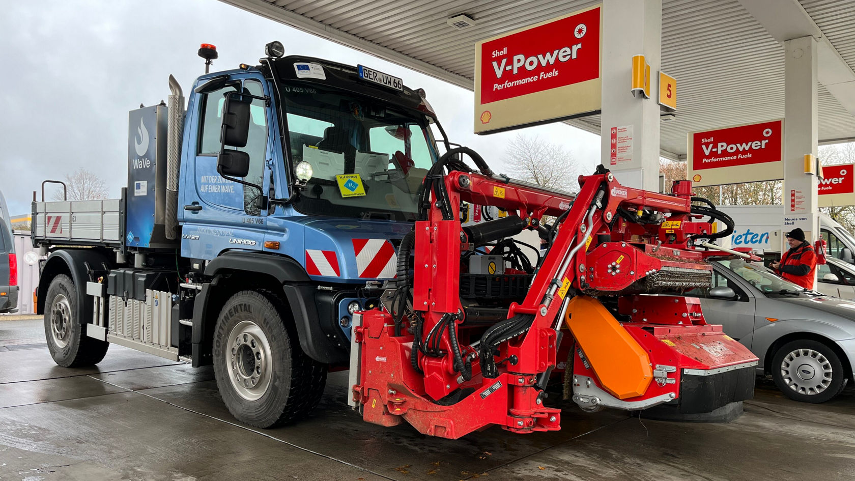 Un Unimog Mercedes‑Benz est stationné dans une station‑service, équipé d'un outil rapporté rouge à l'avant, qui est probablement utilisé pour des travaux agricoles ou communaux.