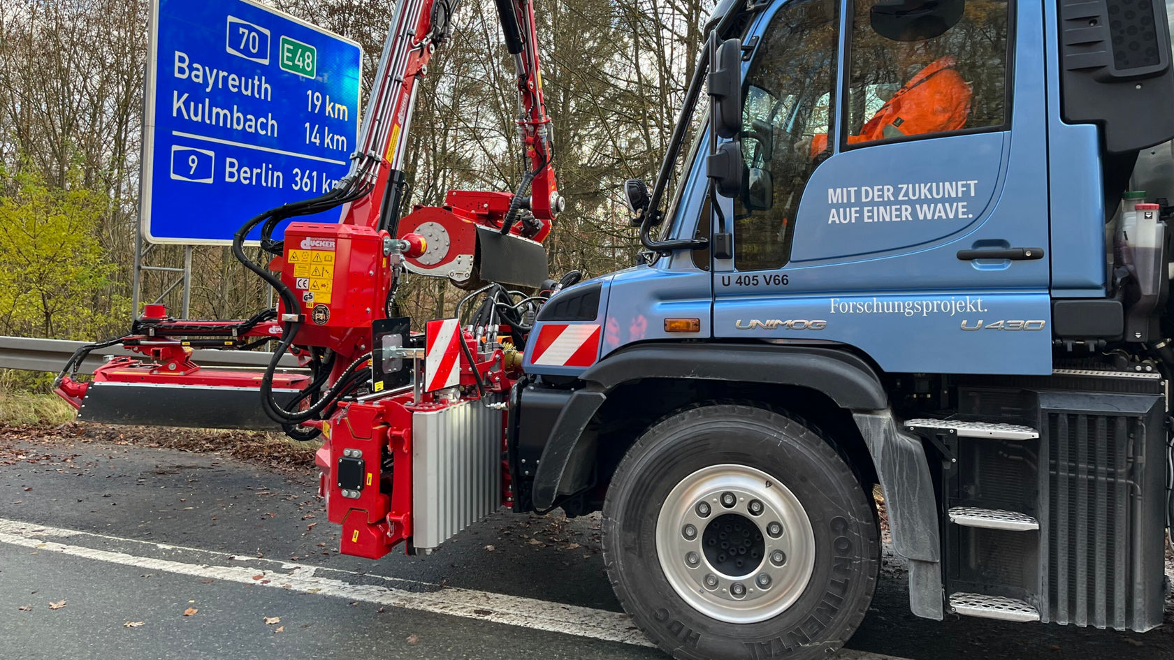 Un Unimog Mercedes‑Benz est stationné au bord de la route, équipé d'un outil rouge pour l'entretien des routes, tandis qu'une forêt est visible en arrière-plan.