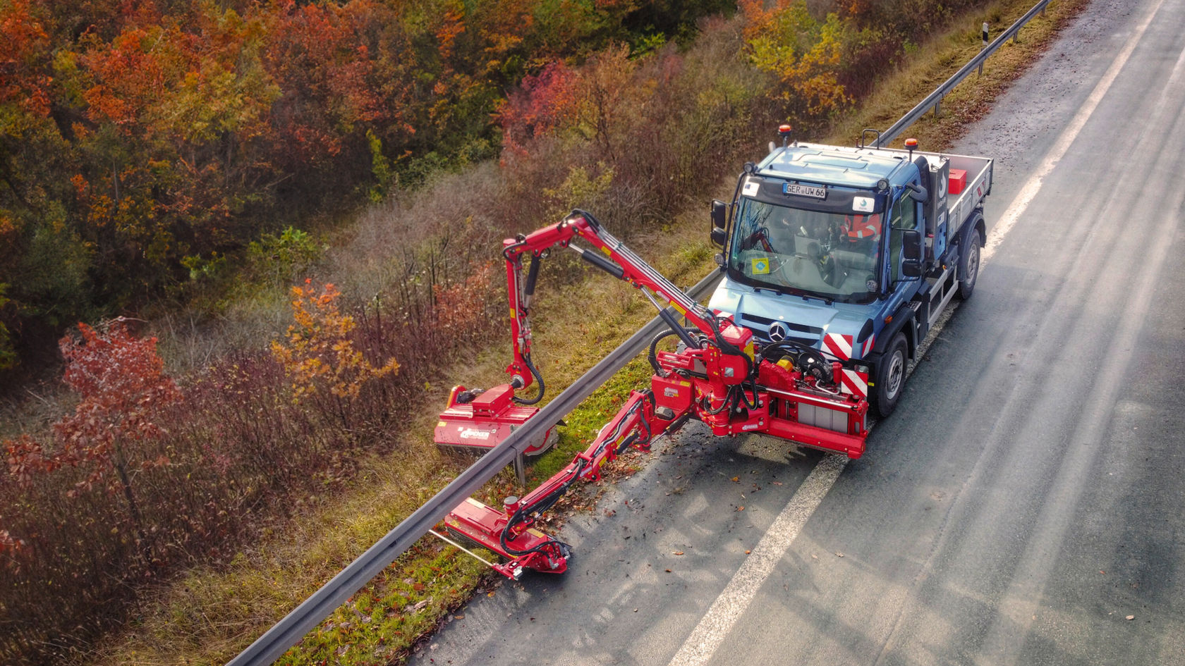 Ein Mercedes-Benz Unimog mit einem Anbaugerät zur Straßenpflege, das entlang einer Straße arbeitet, um die Vegetation zu schneiden. Herbstliche Bäume umgeben den Bereich.