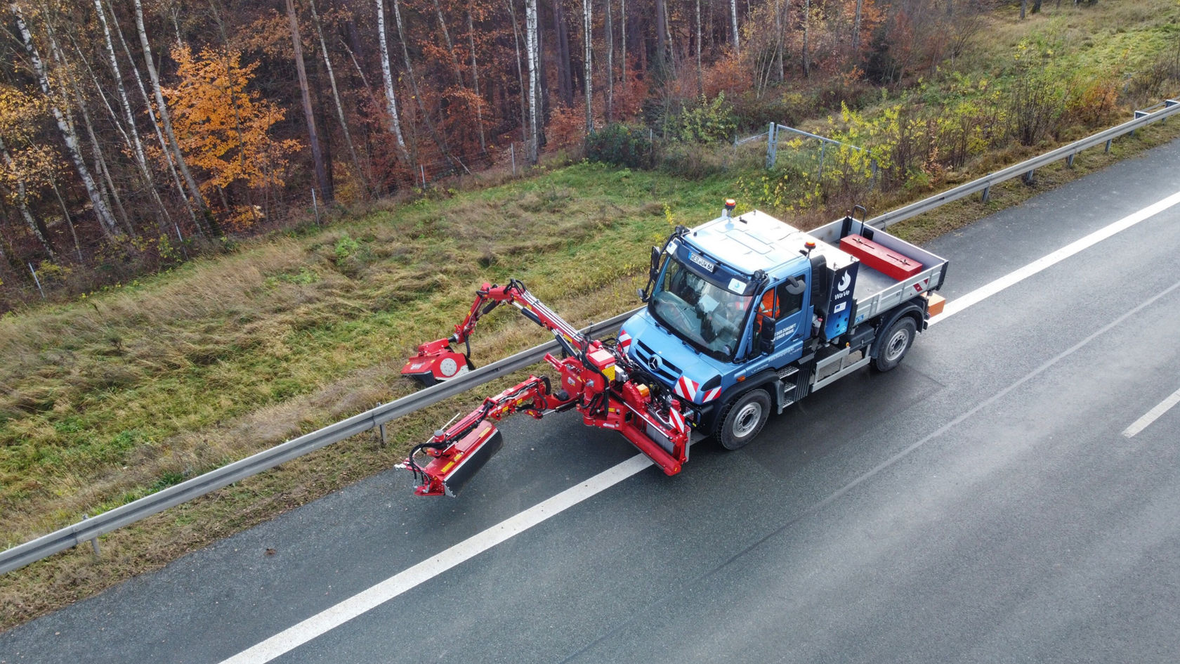 Un Unimog Mercedes‑Benz se trouve sur une route, tandis qu'une faucheuse latérale rouge est déployée sur le côté pour couper de l'herbe ou des mauvaises herbes.