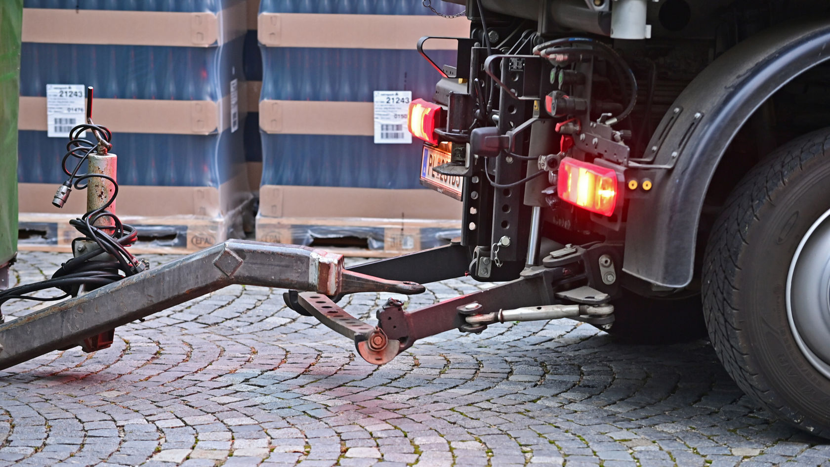 Der hintere Bereich eines Lkw zeigt eine Anhängerkupplung mit Lichtern und Anschlusskabeln, während im Hintergrund gestapelte Container sichtbar sind.
