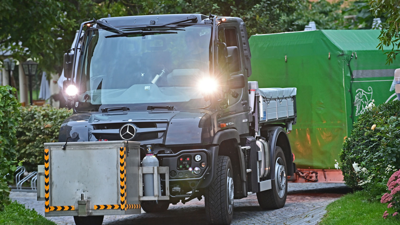 Ein Mercedes-Benz Lkw fährt auf einer gepflasterten Straße, beladen mit einer grünen Plane im Hintergrund. Die Scheinwerfer sind eingeschaltet und der Lkw hat eine robuste Bauweise.