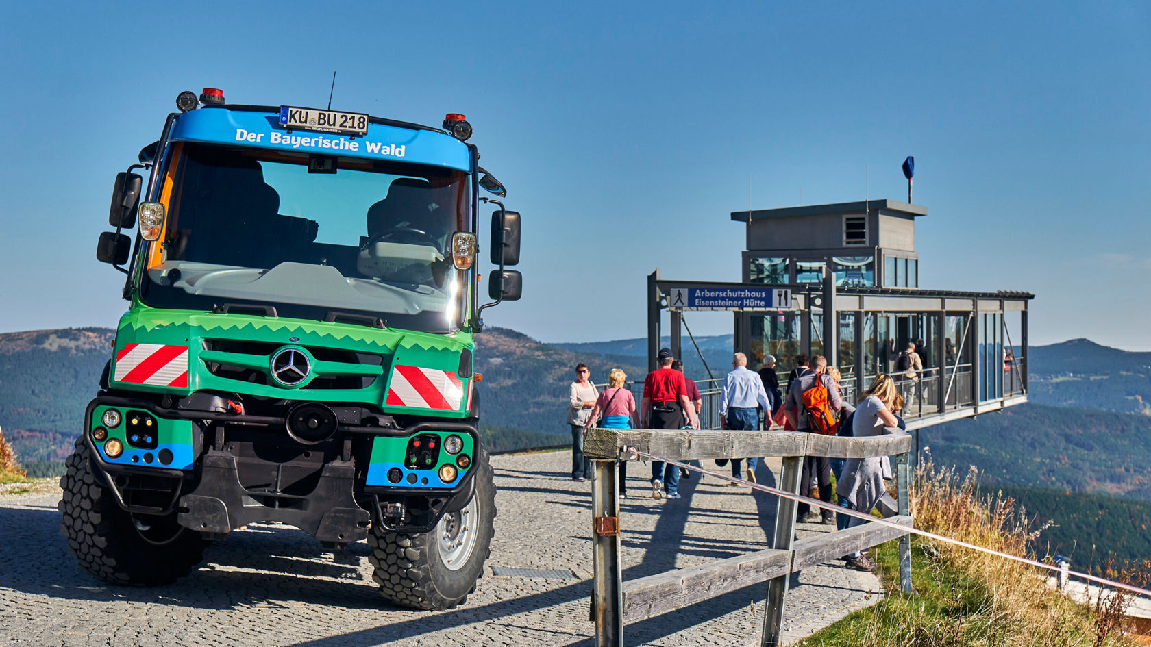 A colorful Mercedes-Benz off-road vehicle stands on a path in the mountains, next to a viewing platform where numerous visitors enjoy the view.