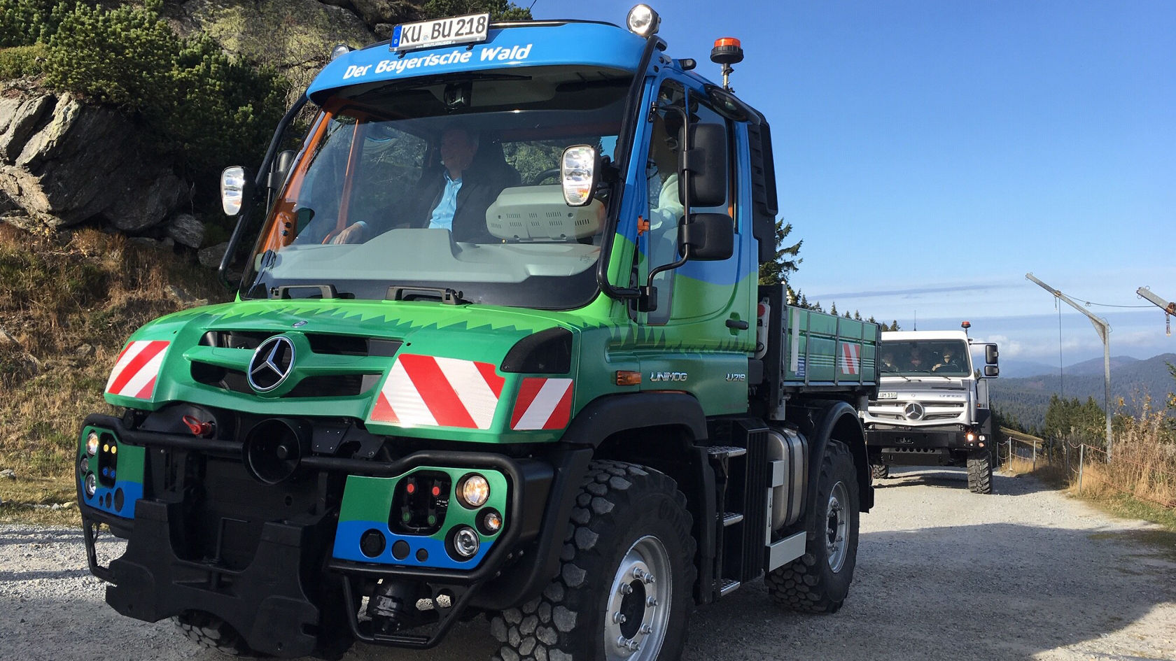 A Mercedes-Benz Unimog in green and blue is driving on a gravel road surrounded by mountain scenery. Another off-roader is visible in the background.