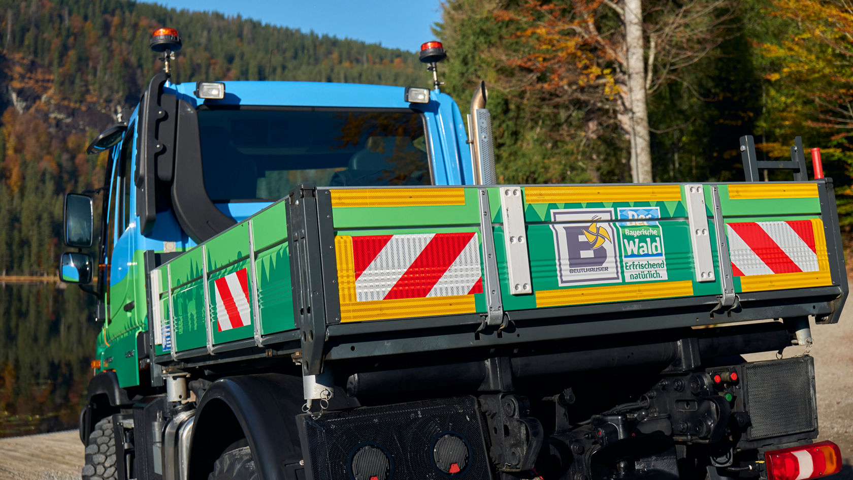 A Mercedes-Benz commercial vehicle with a striking yellow-green loading area stands on the shore of a body of water, surrounded by autumn trees.