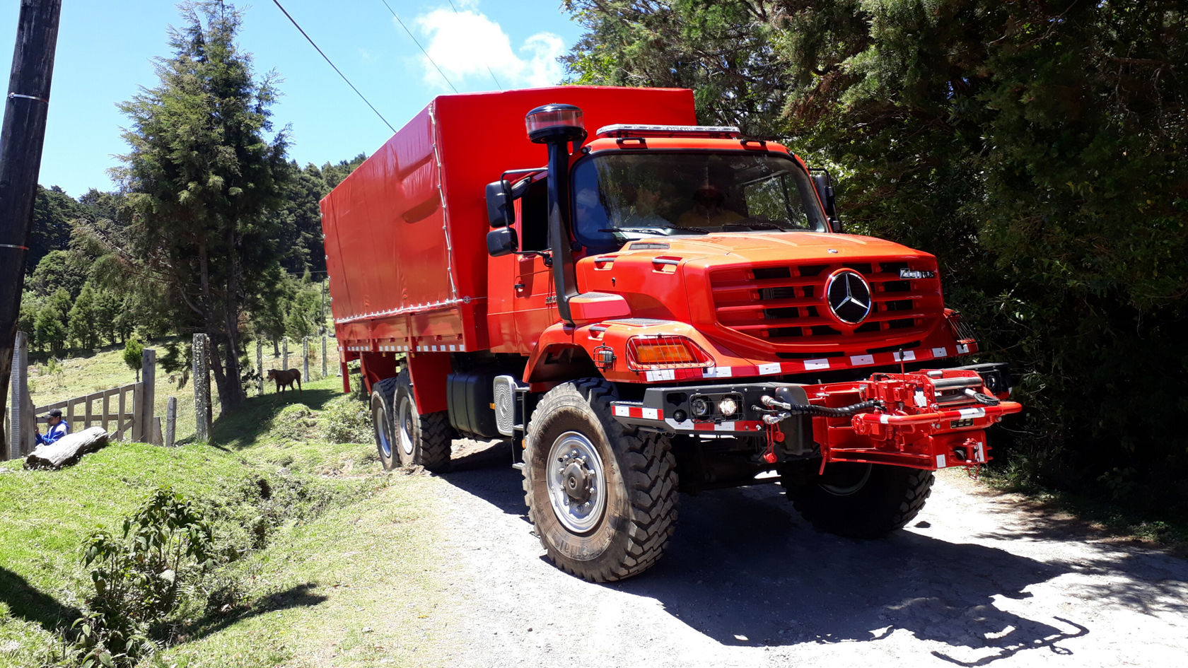Un camion rouge Mercedes‑Benz se trouve sur un chemin de gravier, entouré d'arbres et d'une prairie. Le camion a de grands pneus et une carrosserie robuste.
