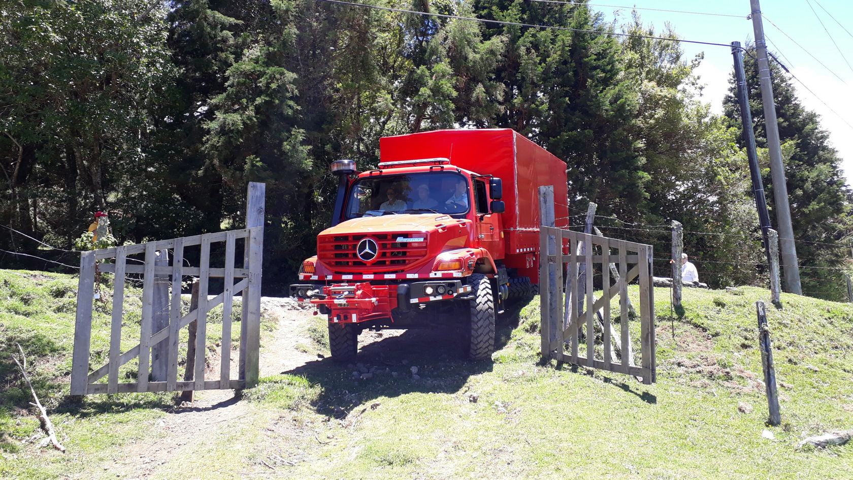 Un camion Mercedes‑Benz rouge franchit un portail ouvert et roule sur un chemin non goudronné, entouré d'une prairie verte et d'arbres.