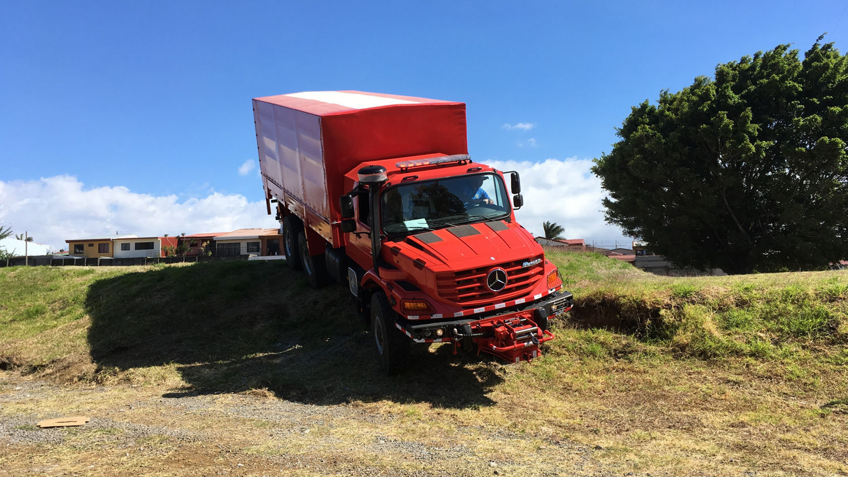 Un camion Mercedes‑Benz rouge est incliné sur une surface inégale, partiellement en pente descendante, avec un compartiment de chargement fermé et un ciel dégagé en arrière-plan.