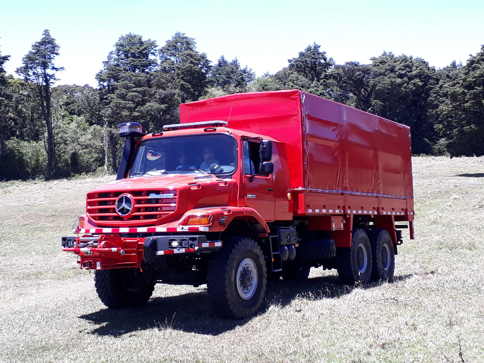 Un camion Mercedes‑Benz rouge doté d'une plate-forme de chargement fermée se trouve sur une prairie verte. Le camion a de gros pneus et une construction robuste, idéal pour le transport de charges lourdes.