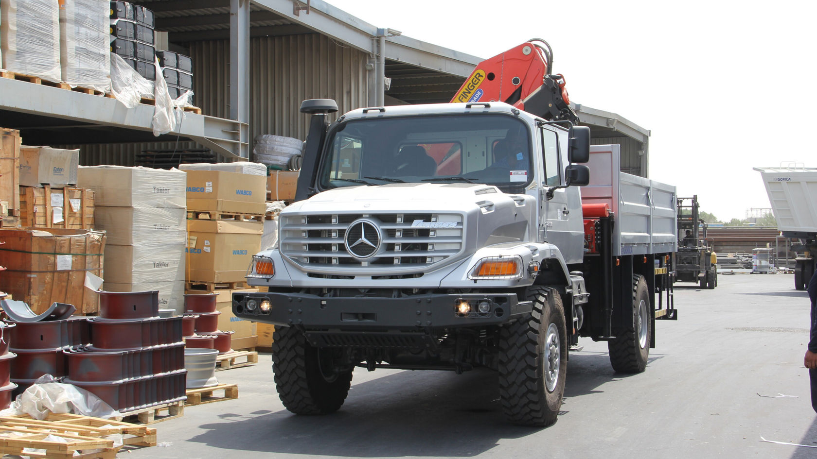 A Mercedes-Benz Zetros with a crane is in a storage area, surrounded by boxes and barrels. The truck is ready to transport goods.