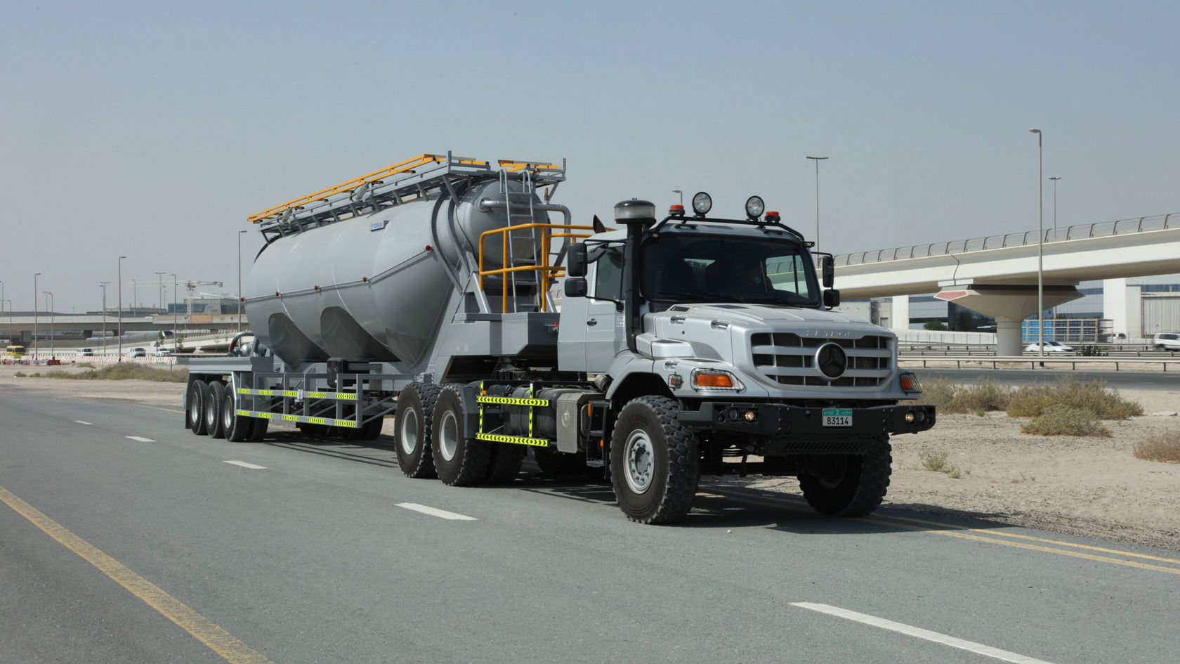 A Mercedes-Benz Zetros is driving on a road and pulling a large, silver tank trailer, surrounded by a dry, urban environment.