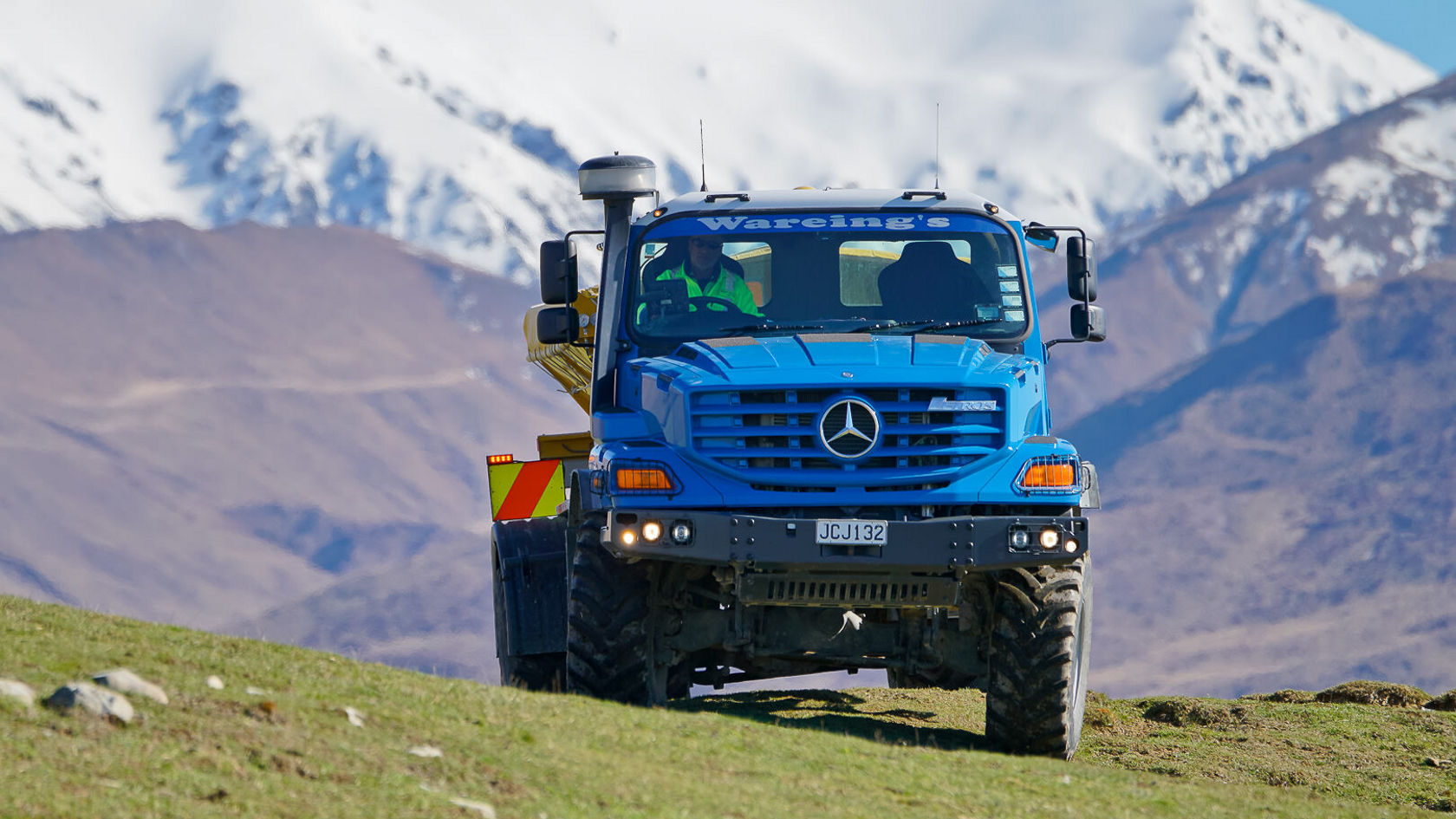 Un camion Mercedes‑Benz bleu roule sur une prairie verte, avec des montagnes enneigées en arrière-plan. Le conducteur est visible et travaille avec un outil rapporté.