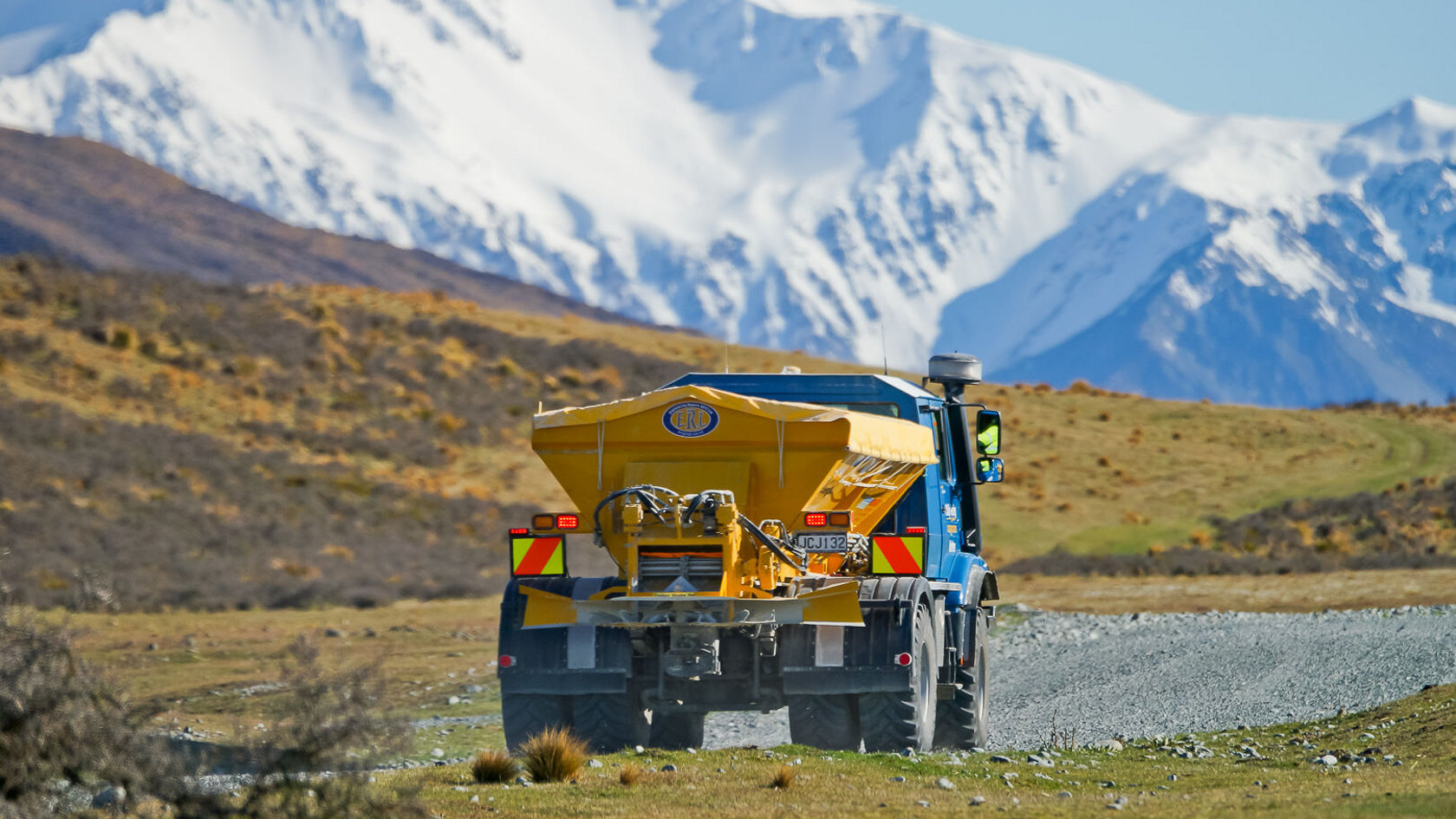 Un camion Mercedes‑Benz bleu avec une semi‑remorque jaune roule sur un chemin de gravier dans un paysage montagneux avec des sommets enneigés.