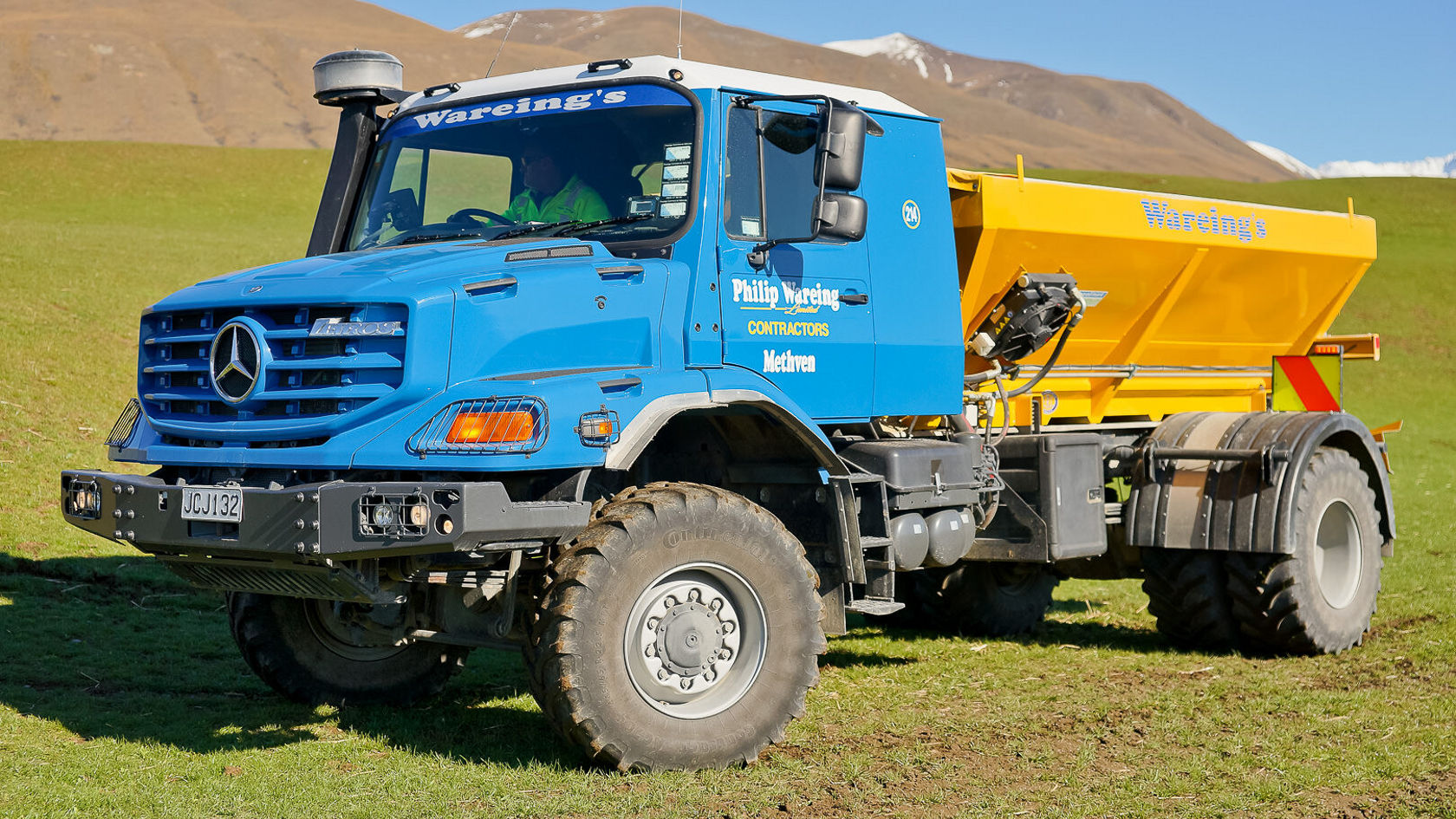 Un camion-benne Mercedes‑Benz bleu avec une benne jaune se trouve sur une prairie verte, utilisé pour des travaux de construction, doté de pneus robustes et de détails modernes.