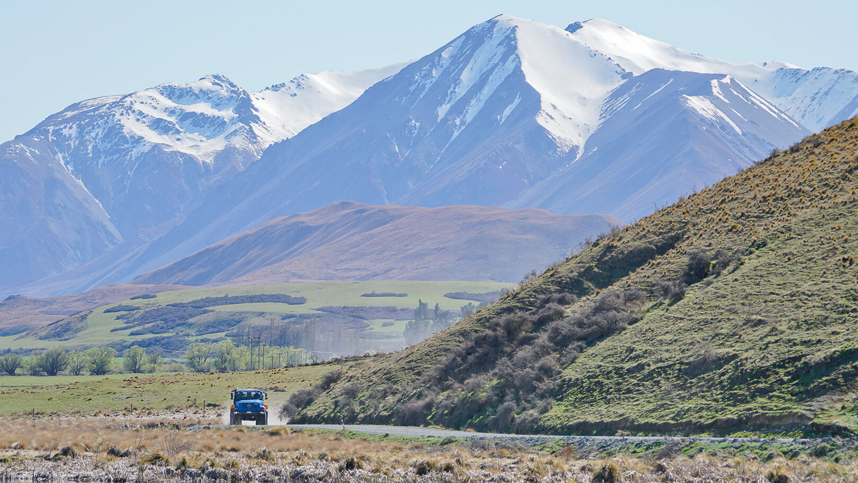 Un véhicule Mercedes‑Benz circule sur une route sinueuse longeant un plan d'eau paisible, entouré de collines verdoyantes et de montagnes majestueuses en arrière-plan.