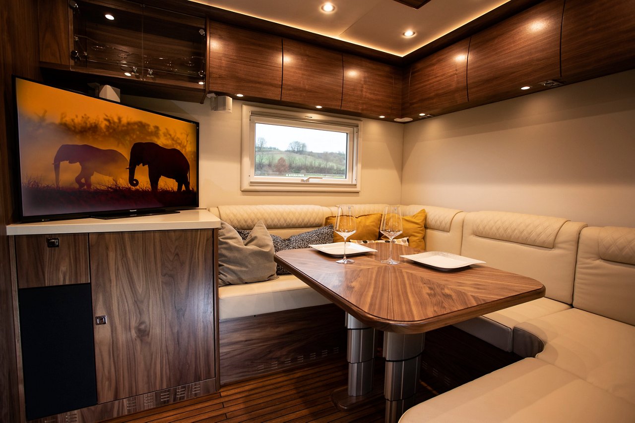 Ein moderner Wohnraum in einem Zetros Wohnmobil mit einem Holztisch, bequemen Sitzpolstern und Fernseher. Die Fenster bieten einen Blick auf die Umgebung.