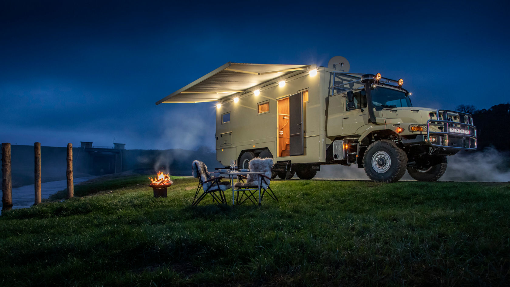 Ein Zetros Wohnmobil steht auf einer Wiese bei Nacht. Das Fahrzeug hat ein beleuchtetes Ausklappdach. Im Vordergrund sind zwei Stühle und ein kleines Feuer sichtbar.