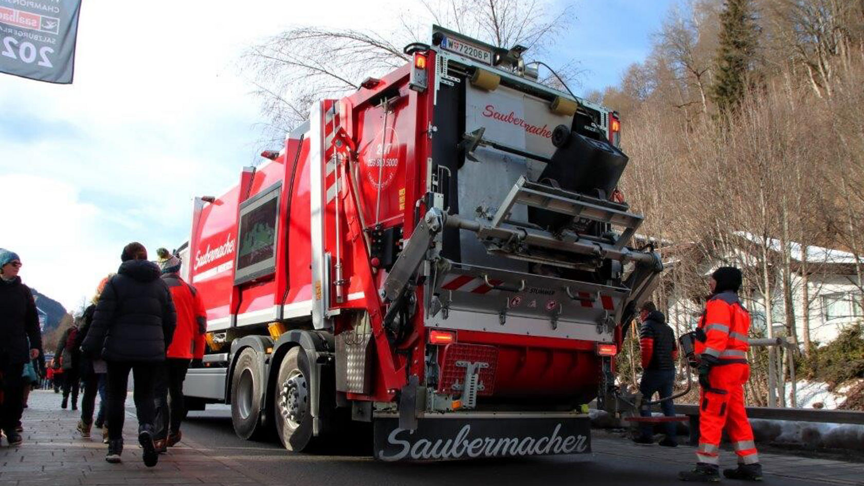 A red Mercedes-Benz refuse truck is parked on a street, while several people in work clothes stand nearby and move about.
