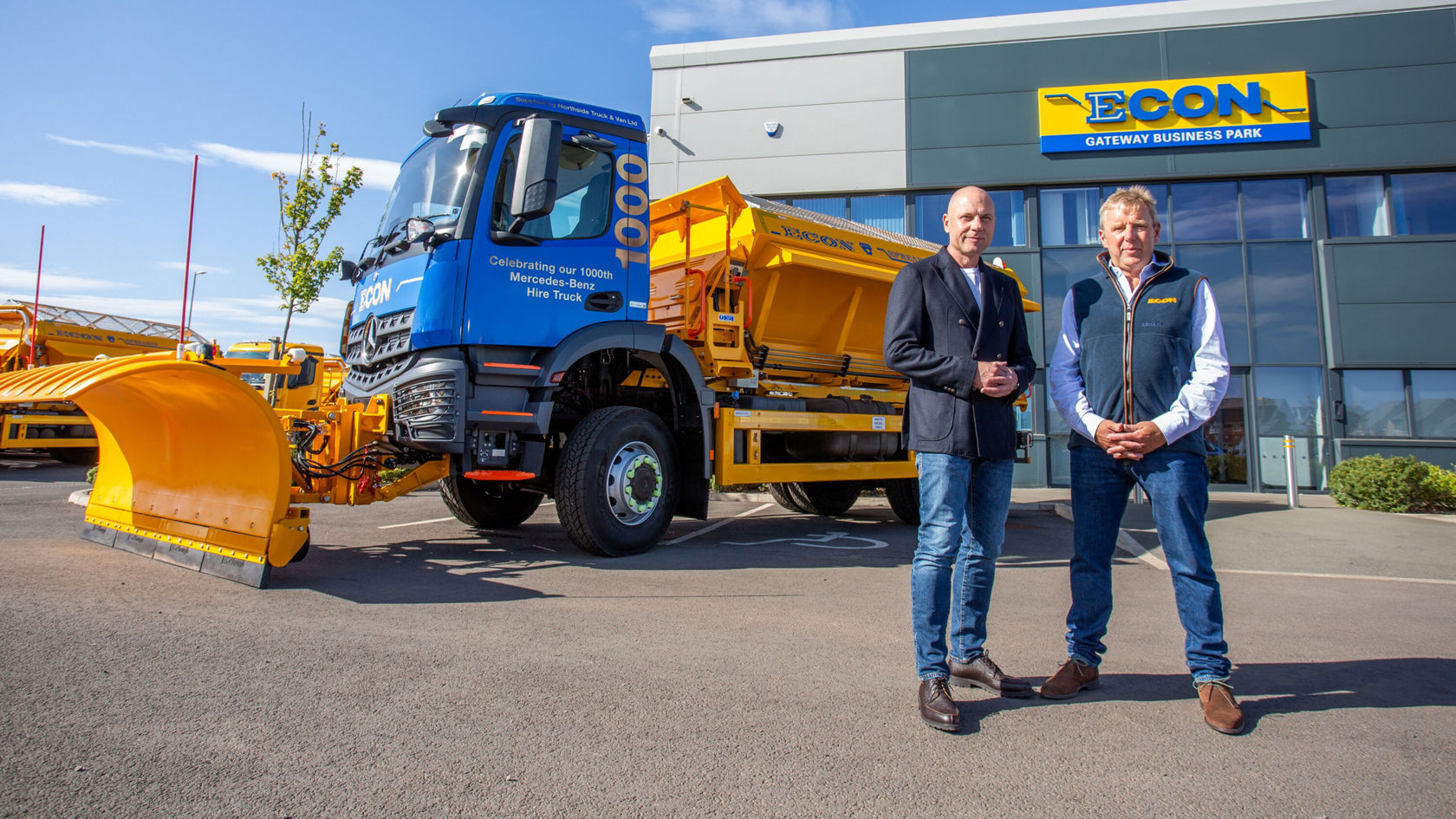 Two men are standing next to a Mercedes-Benz Arocs truck with a yellow snowplow and a flatbed. In the background, the building of the ECON Gateway Business Park is visible.