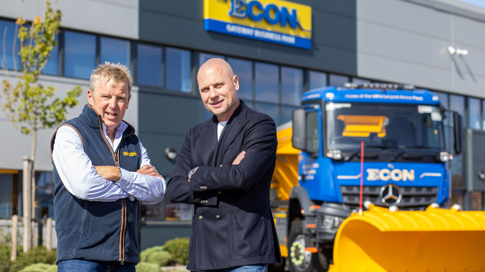 Two men are standing, smiling, in front of a Mercedes‑Benz Arocs with a snowplough, near the ECON building, which is marked with a large logo.