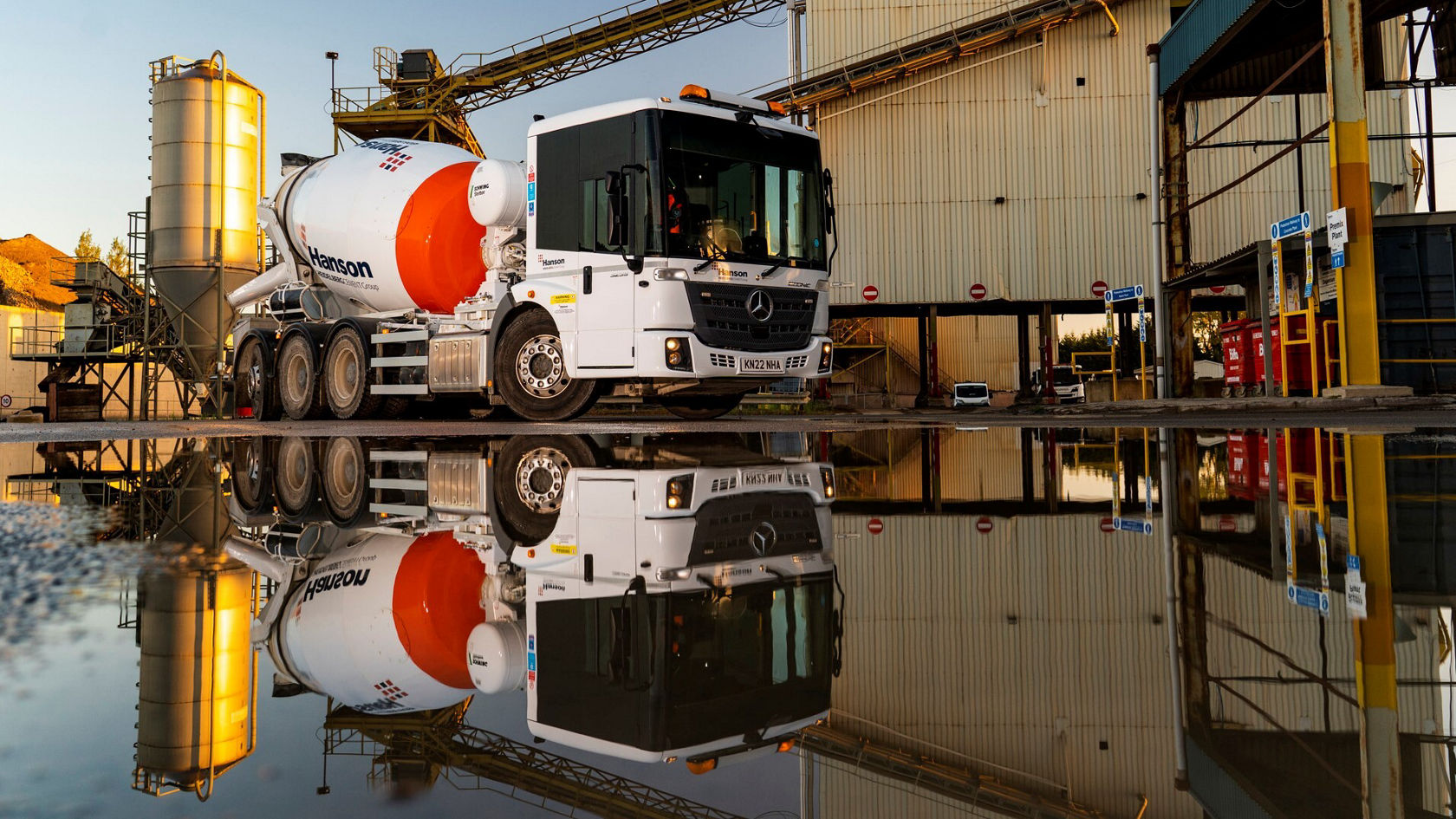 A Mercedes-Benz Econic concrete mixer stands in front of a concrete plant, reflected in a puddle. The scene shows construction activities with a focus on powerful vehicle technology.