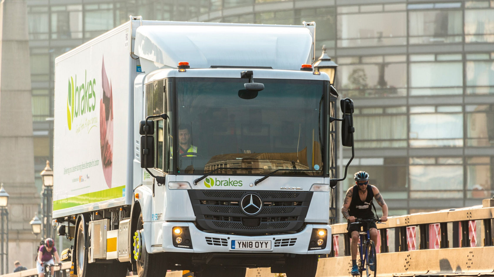 Un camion Econic Mercedes-Benz circule sur un pont, alors qu'un cycliste roule à côté de lui. Le camion porte le logo de la marque Brakes et transporte des marchandises.