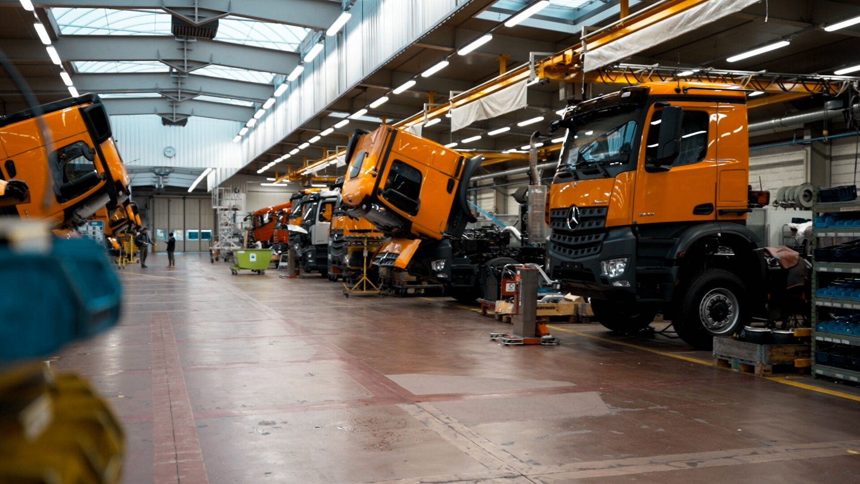 In a workshop, several orange Mercedes-Benz Econic trucks can be seen at various stages of assembly while mechanics work on the vehicles.