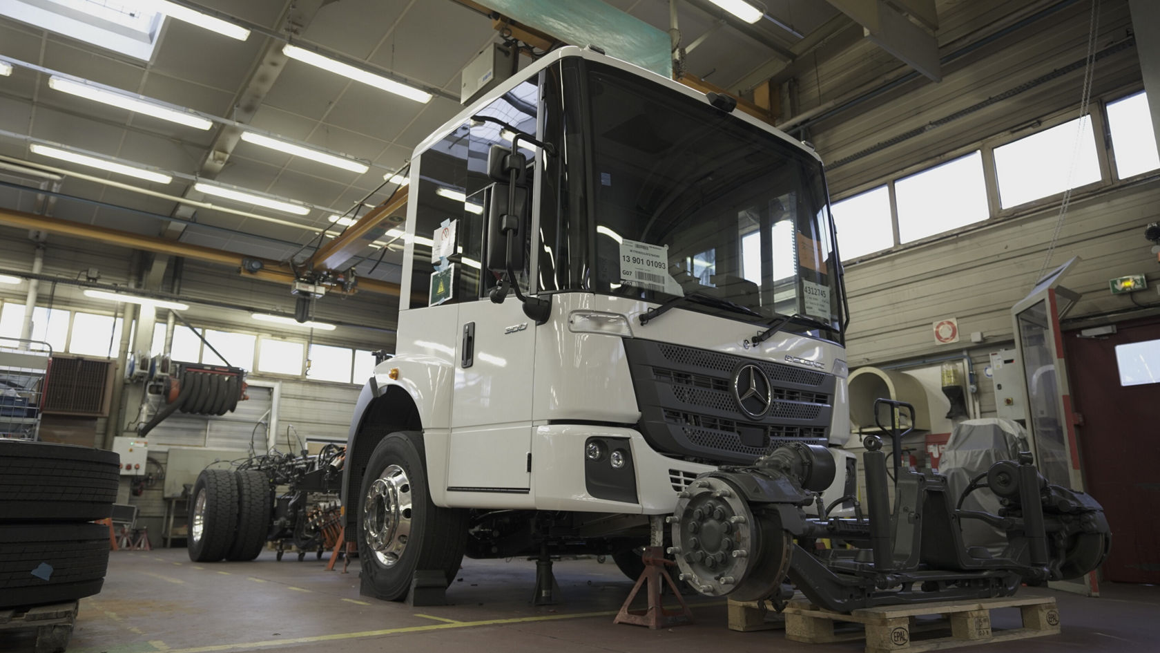 A Mercedes-Benz Econic truck is in a workshop, partially disassembled, with the chassis and various vehicle components visible in the background.