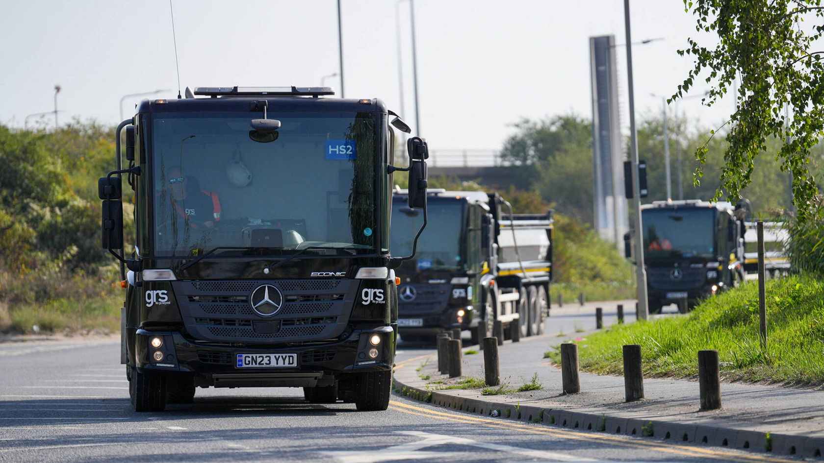 A Mercedes-Benz truck is driving on a road, followed by other trucks of the same brand. The scene shows an urban environment with few traffic signs.