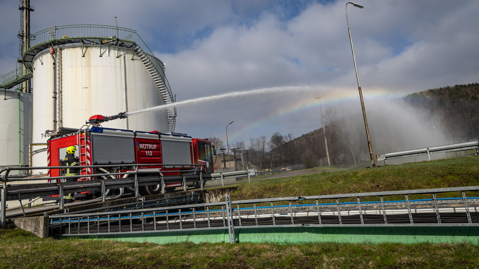 Un véhicule de pompiers projette de l'eau dans les airs, tandis qu'en arrière-plan une cuve de stockage et un arc‑en‑ciel sont visibles. La scène montre un exercice d'entraînement à la lutte contre les incendies.