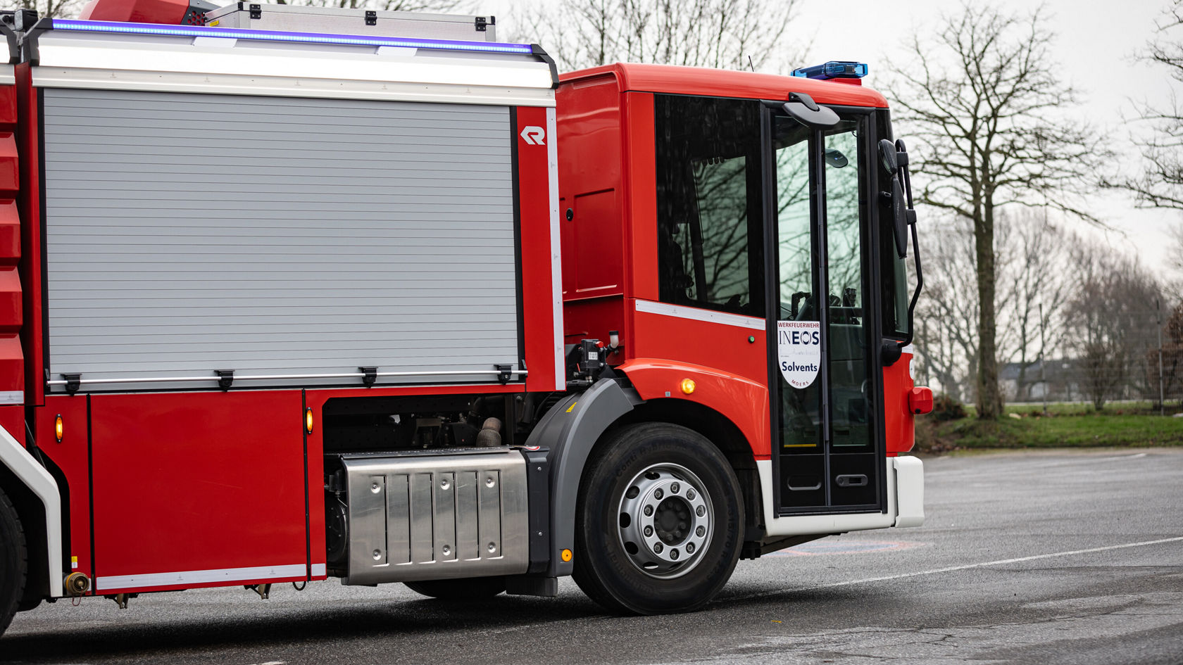 Un véhicule d'incendie rouge avec une grande porte latérale coulissante est stationné sur un parking, tandis que des arbres sont visibles en arrière-plan. Le véhicule est équipé d'un gyrophare bleu sur le toit.