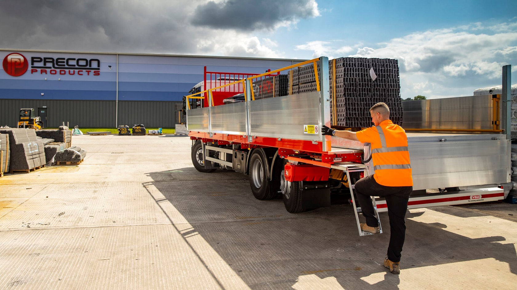A Mercedes-Benz Econic truck is parked in a parking lot while a worker in an orange safety vest unloads materials from the truck in front of a building with the sign "Precon Products".