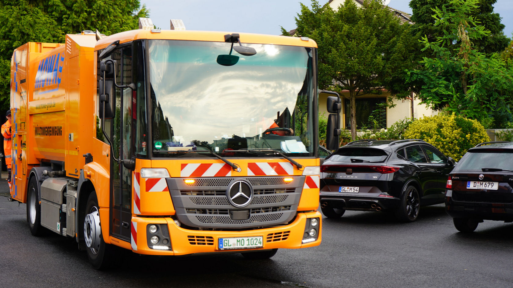 Een oranje Mercedes-Benz Econic vuilniswagen staat op een straat, omgeven door geparkeerde auto's. De voorkant toont waarschuwingsmarkeringen en het kenteken is zichtbaar.