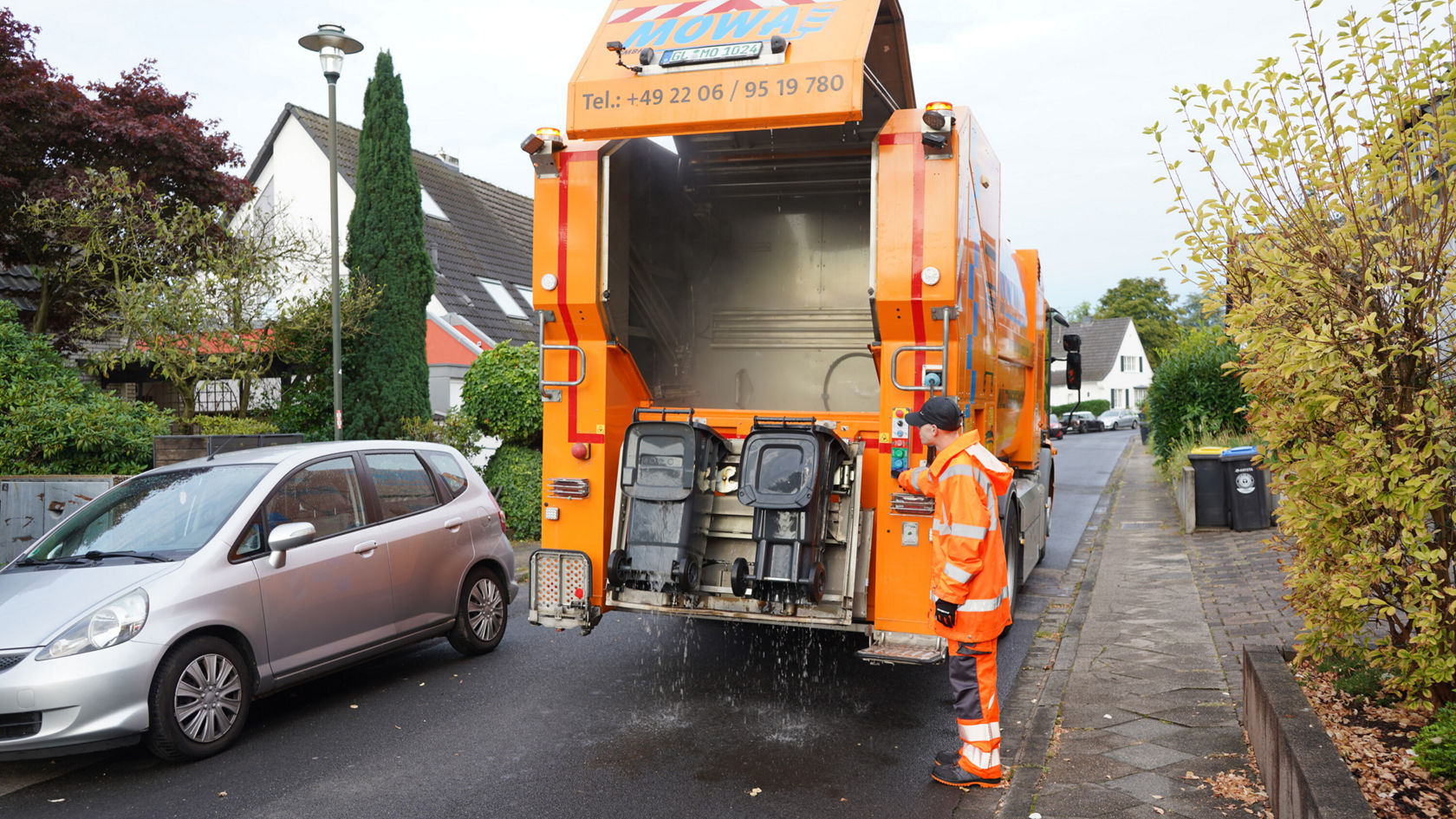 Een oranje Mercedes-Benz Econic vuilniswagen leegt zijn inhoud, terwijl een medewerker in uniform toezicht houdt op de handeling. Woonhuizen en een geparkeerde auto zijn op de achtergrond zichtbaar.