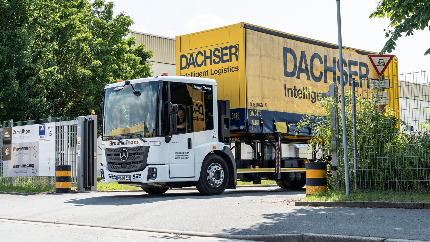 Ein Mercedes-Benz Econic-Lkw fährt durch das Tor eines Logistikzentrums, beladen mit einem gelben Dachser-Container, umgeben von Bäumen und einer Lagerhalle.