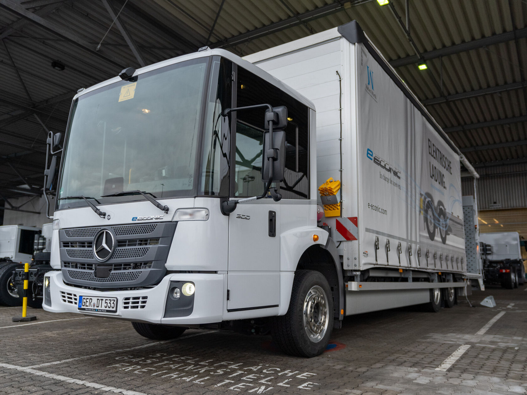 Ein Mercedes-Benz Econic Lkw steht in einer Lagerhalle, ausgestattet mit einem Auflieger. Der Lkw ist weiß mit einem großen Unternehmenslogo und Informationen über Elektrofahrzeuge.