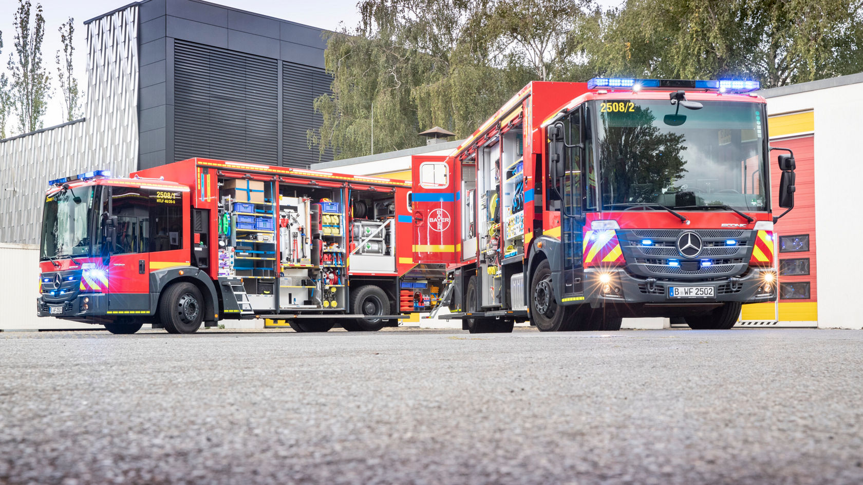 Zwei Feuerwehrfahrzeuge von Mercedes-Benz stehen vor einem Feuerwehrgebäude. Eines der Fahrzeuge ist mit einer offenen Ladefläche für Ausrüstung versehen, während das andere bereit für den Einsatz ist.