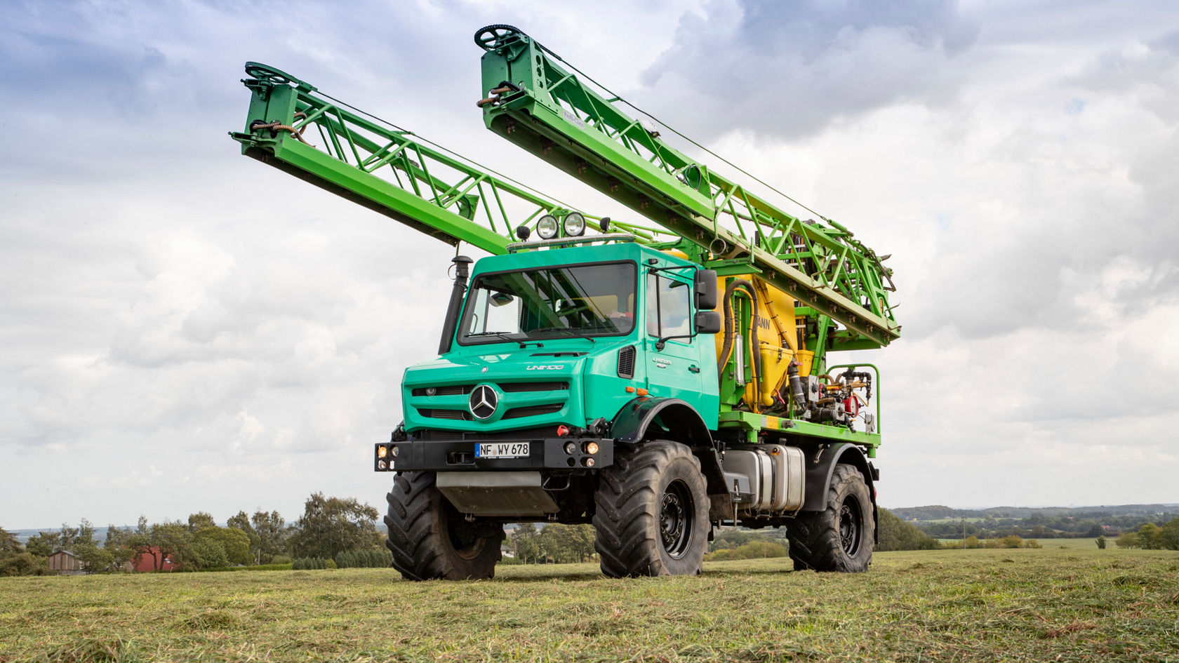 Ein grüner Mercedes-Benz Unimog mit einem Hubgerüst steht auf einem Grasfeld. Der Traktor ist für landwirtschaftliche Aktivitäten konzipiert und zeigt robuste Räder und eine gelbe Aufbaukonstruktion.