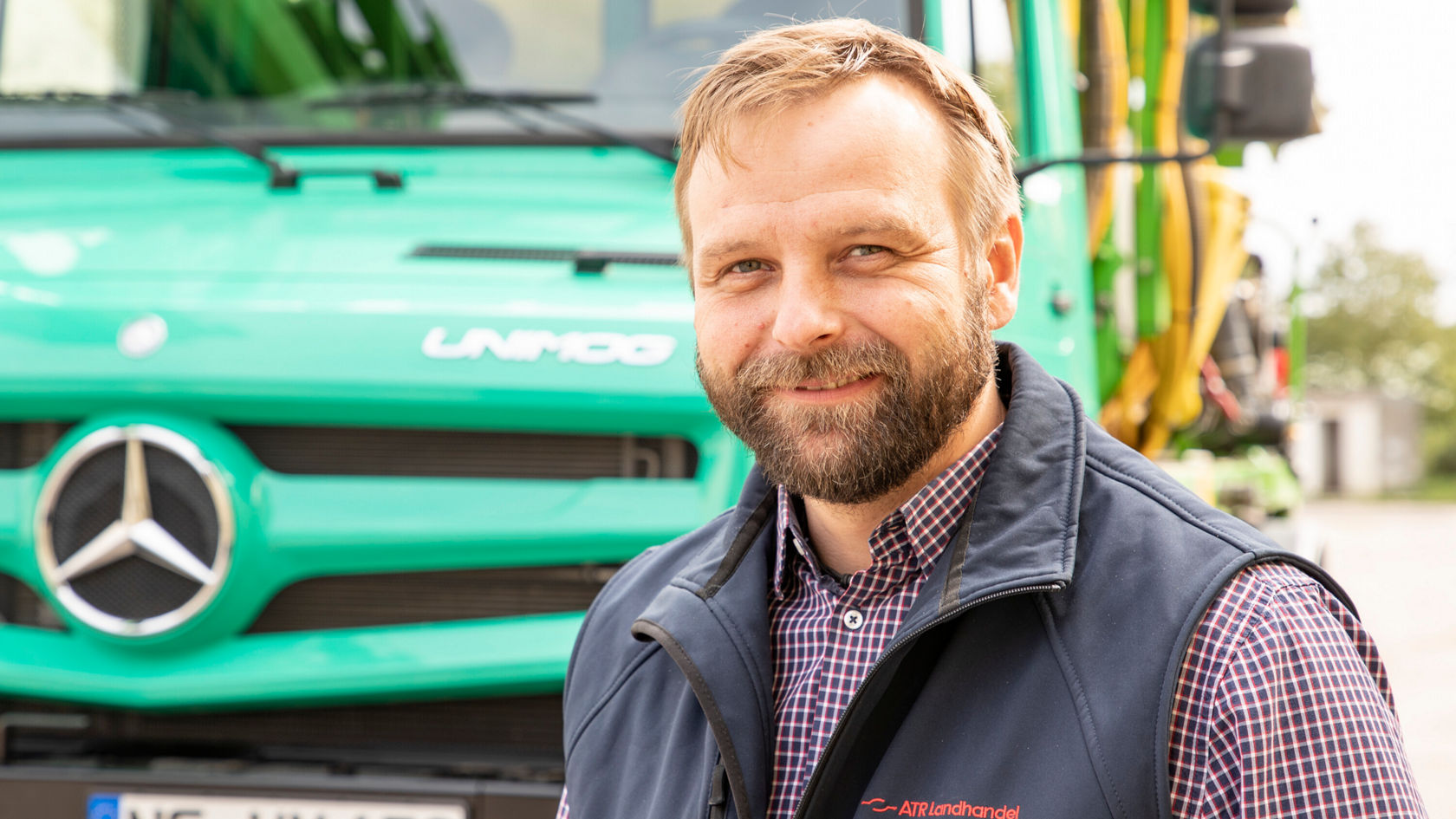 Ein Mitarbeiter steht lächelnd vor einem grünen Mercedes-Benz Lkw, gekleidet in einem dunklen Weste und einem karierten Hemd.