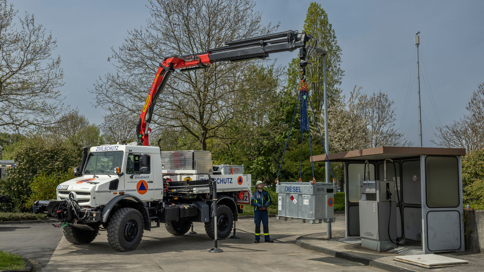 A Mercedes-Benz Unimog with a crane is lifting a load at a petrol station while a person in work clothing watches in close-up. Trees and an adjacent station are visible in the background.