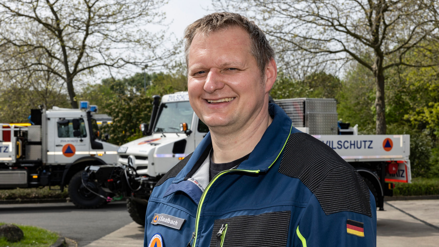 A smiling man in work clothes stands in the foreground, while two disaster relief vehicles are visible in the background.