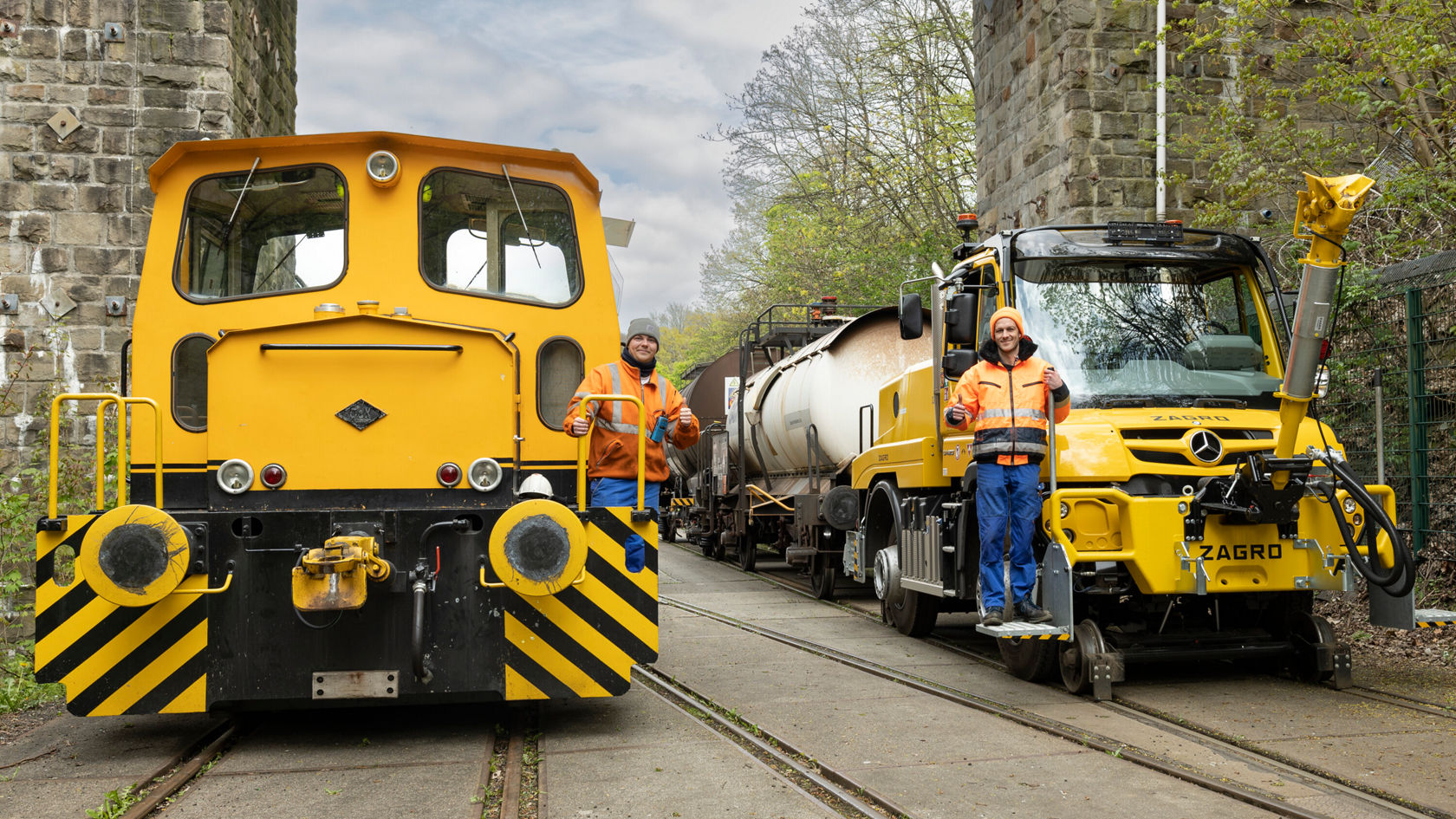 Zwei Arbeiter stehen neben einer gelben Lokomotive und einem Mercedes-Benz Unimog auf einem Gleis. Im Hintergrund sind Bäume und eine Steinmauer sichtbar.
