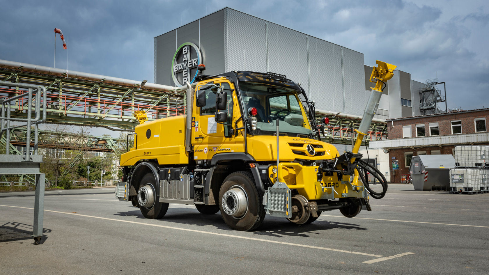 Ein gelber Mercedes-Benz Unimog steht auf einem Industriegelände. Er ist mit einer Arbeitsvorrichtung und einer großen Frontschaufel ausgestattet.