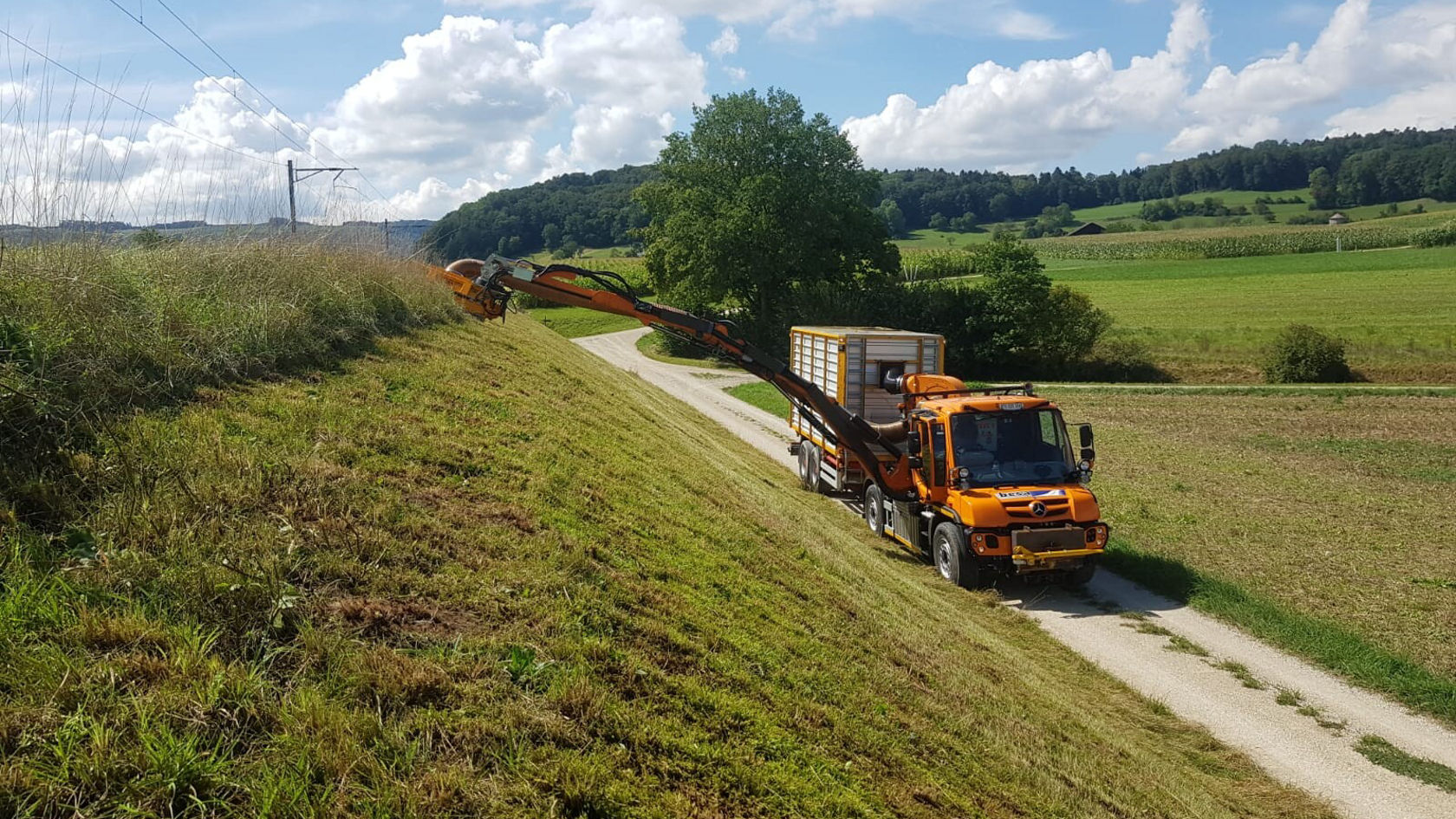 An orange Mercedes-Benz mower is cutting grass along a slope beside a narrow path, surrounded by fields and trees under a blue sky. An orange Mercedes-Benz mower is cutting grass along a slope beside a narrow path, surrounded by fields and trees under a blue sky.