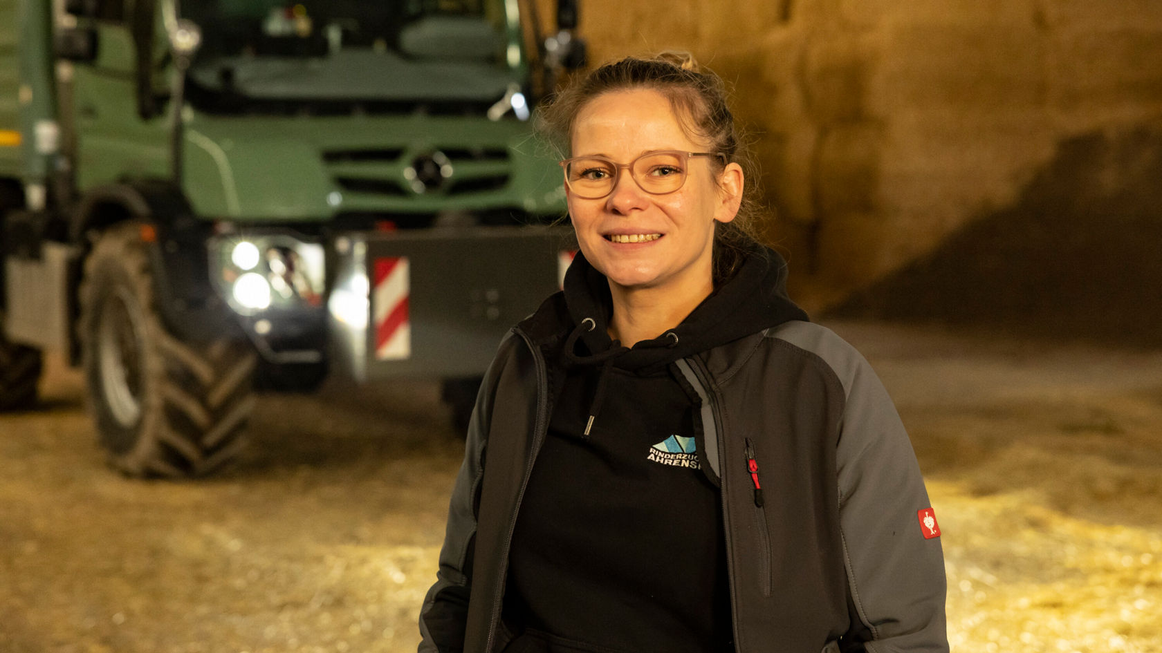 Eine Person mit Brille und einem grauen Mantel lächelt in einer Scheune, während im Hintergrund ein Mercedes-Benz Unimog zu sehen ist.