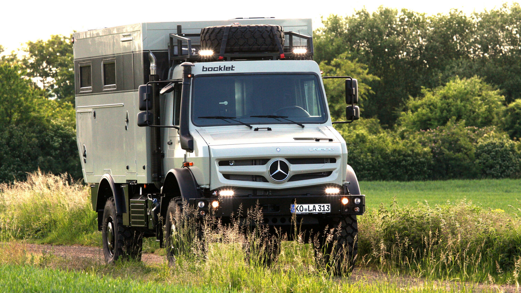 A Mercedes-Benz Unimog is driving along a field track, surrounded by tall grasses. The vehicle is equipped with an attachment for camping equipment and has additional headlamps.