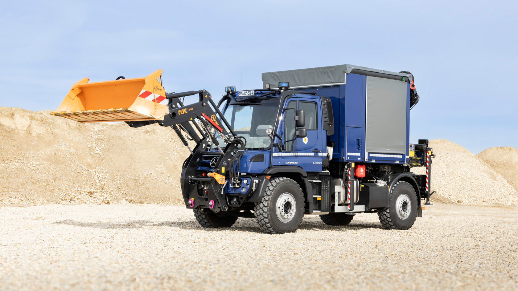 Un Unimog Mercedes‑Benz bleu, équipé d'un bras de chargeuse déployé, est stationné sur un terrain en gravier.
