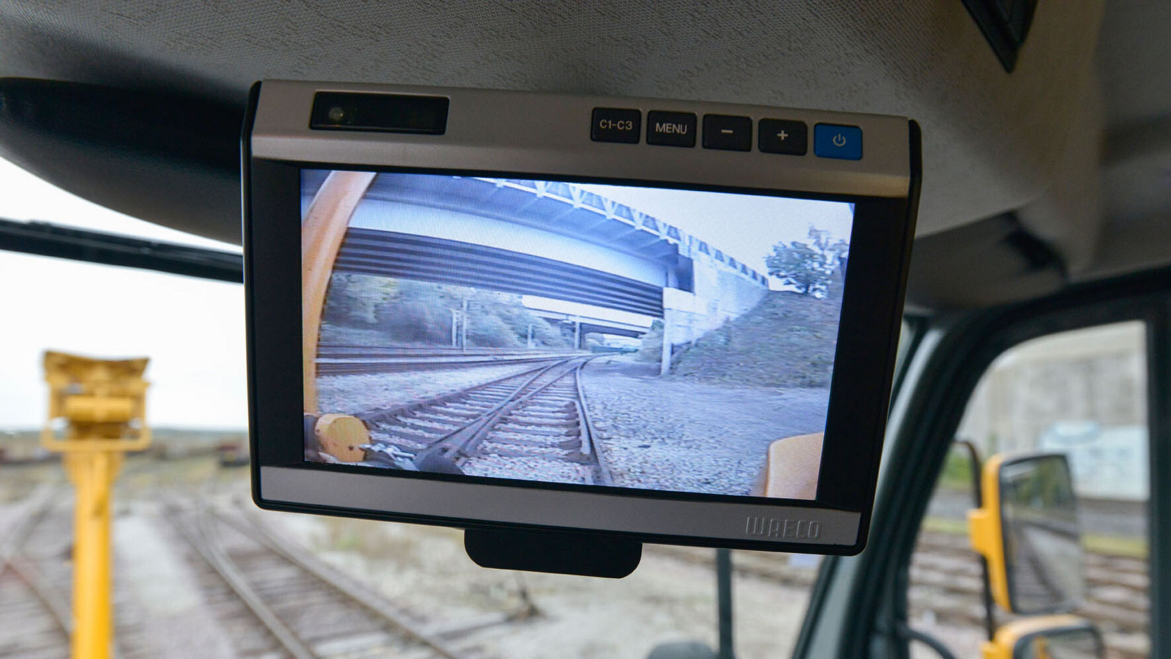 Ein Monitor in einem Fahrerkabine zeigt eine Perspektive auf Gleise unter einer Brücke. Der Zustand der Gleise und die Brücke sind deutlich sichtbar.